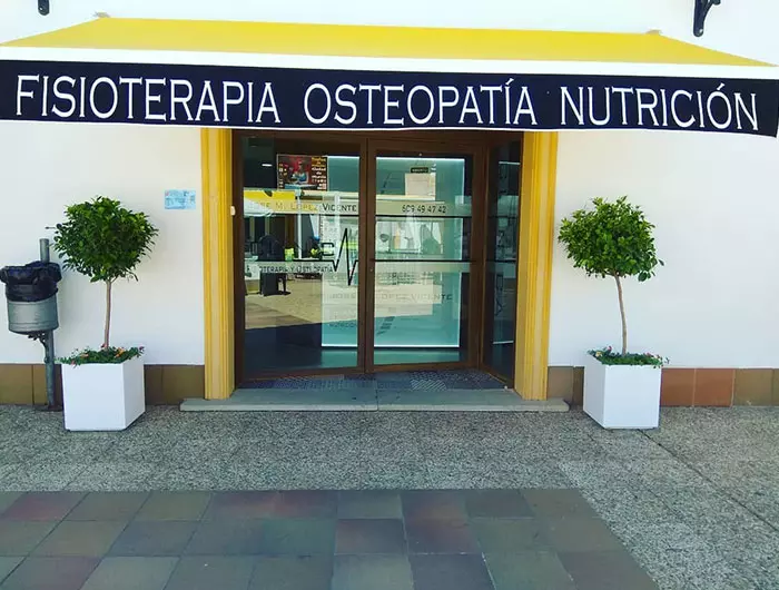 Fachada de Clínica La Alcayna con toldo amarillo, entrada de cristal y rótulos de Fisioterapia, Osteopatía y Nutrición